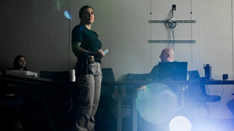 Female cop standing next to a male cop sitting at a computer, posed to depict weapons training.
