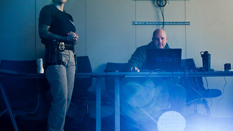 Female cop standing next to a male cop sitting at a computer, posed to depict weapons training.