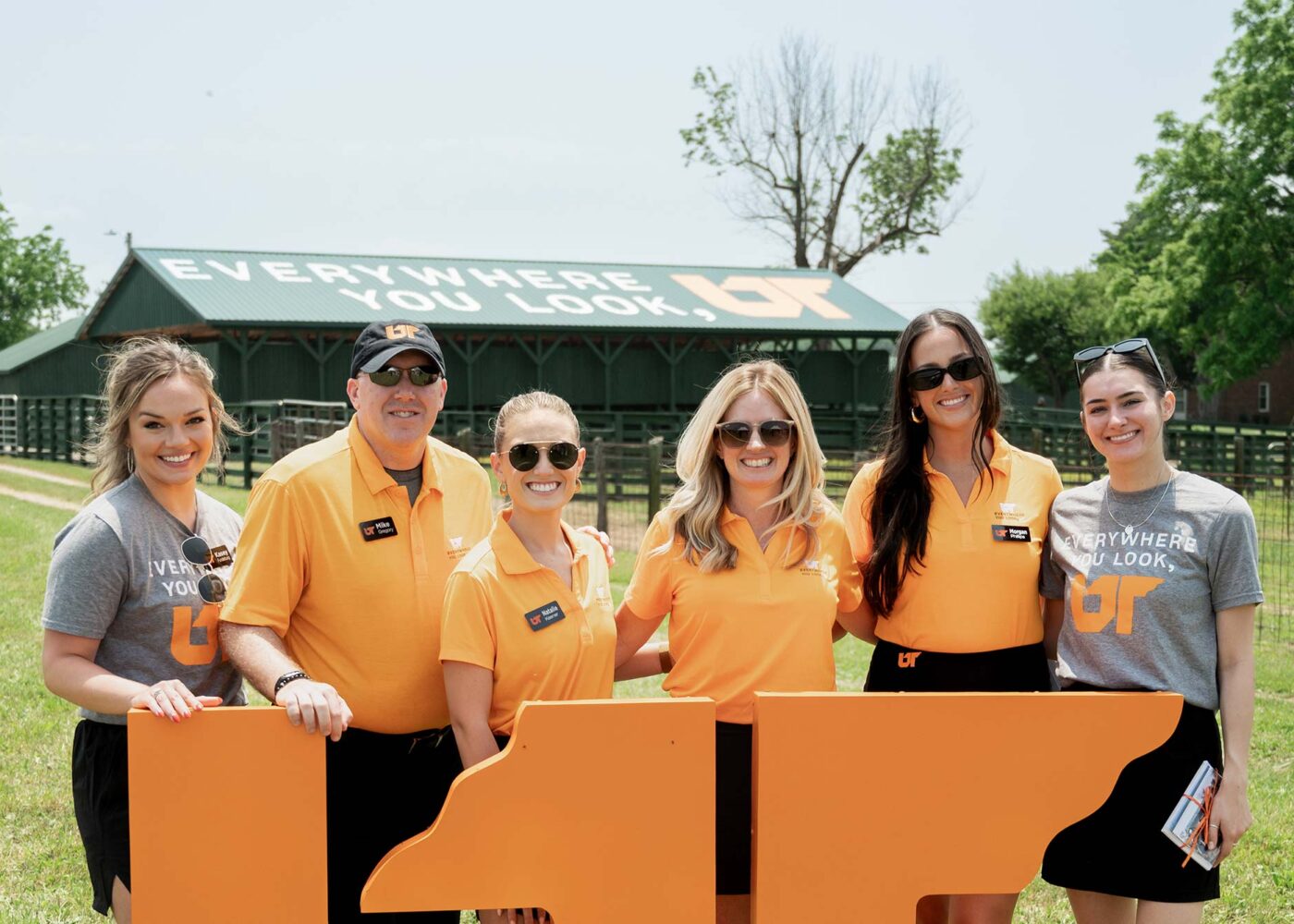 A group of people wearing orange and gray shirts smile for a photo in an open field, with a green-roofed barn behind them.