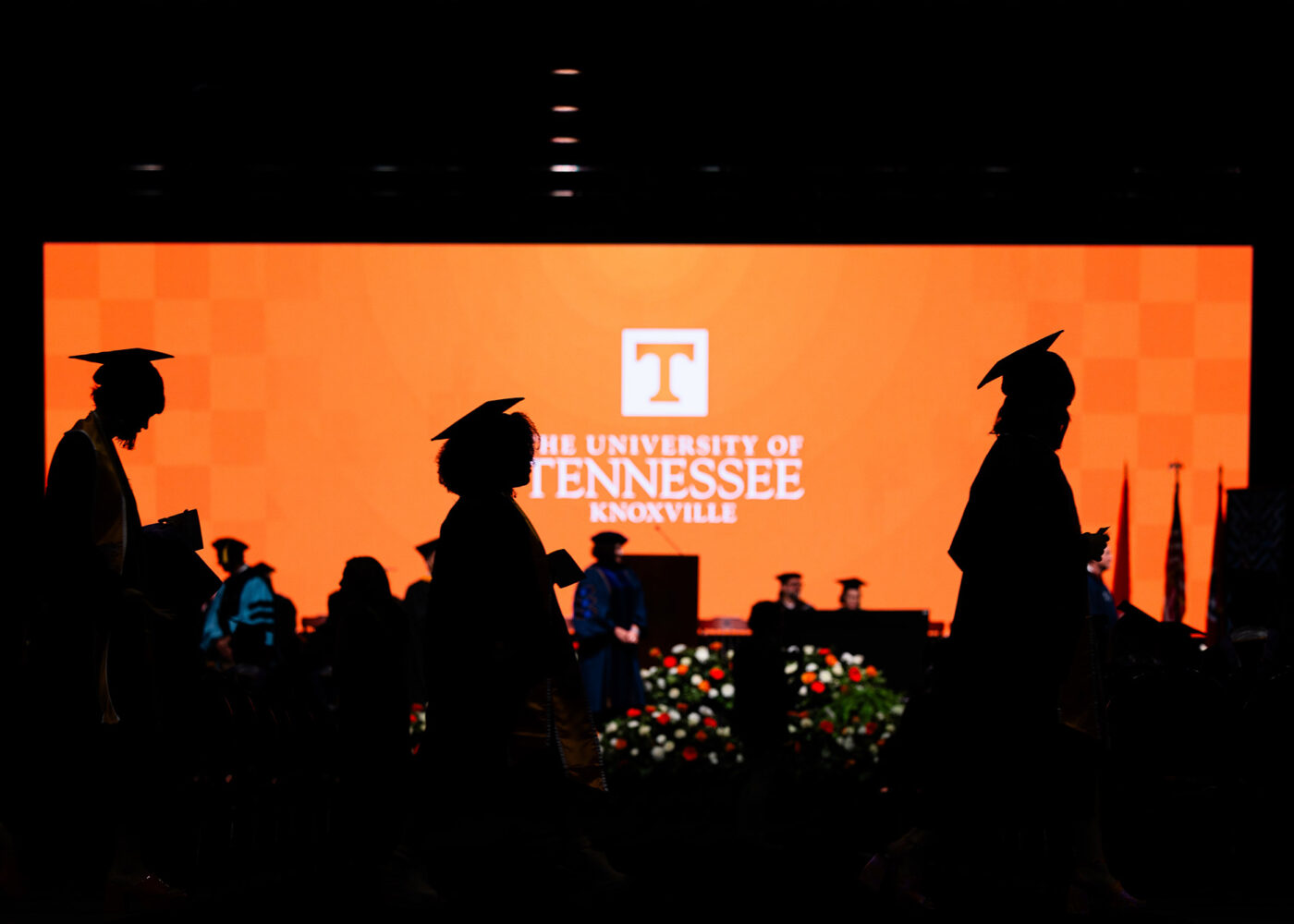 Silhouettes of students walking across graduation stage, lit by a bright orange screen in the background.
