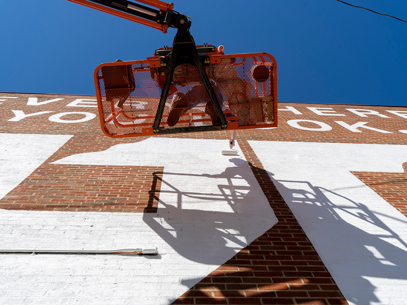 An artist uses a lift to paint a large UT mural on the side of a building.