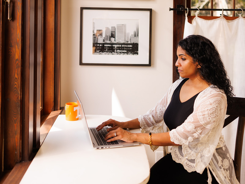 A woman sits at a white table, using her laptop.
