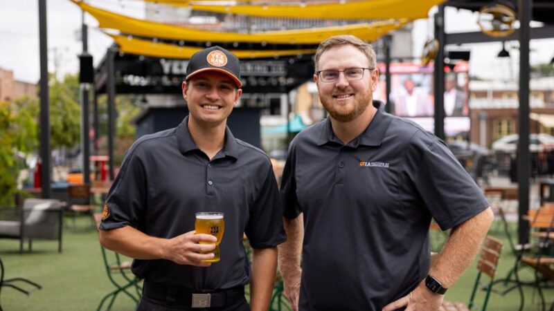 Two men wearing gray polo shirts smile for a photo while holding a glass of beer.