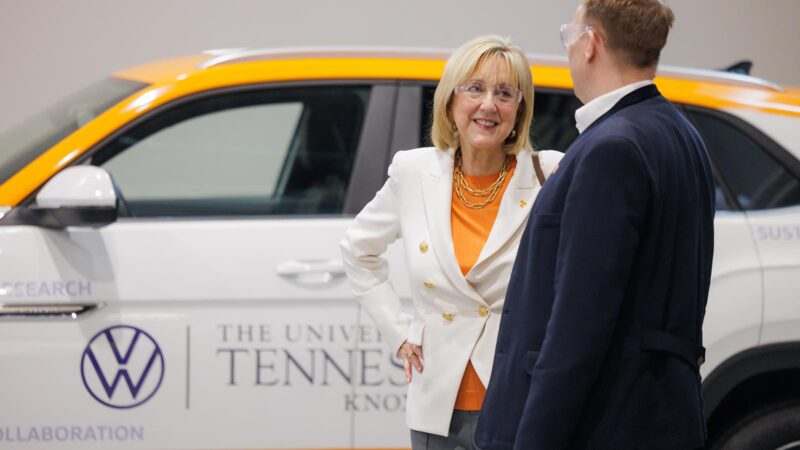 A man and woman, both wearing safety glasses, talk while next to a white Volkswagen vehicle.