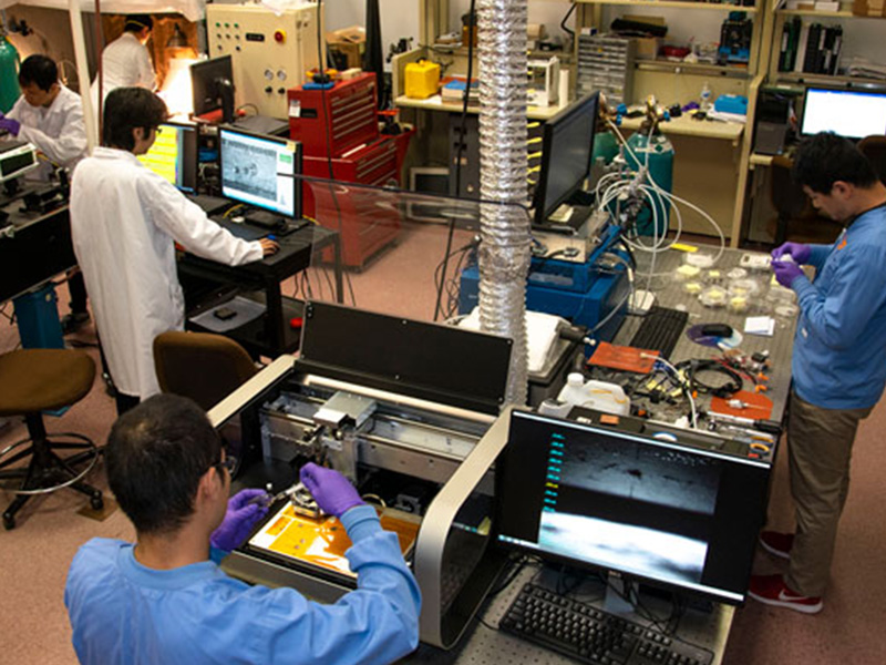 Three men wearing lab coats work in a research laboratory on various pieces of equipment.