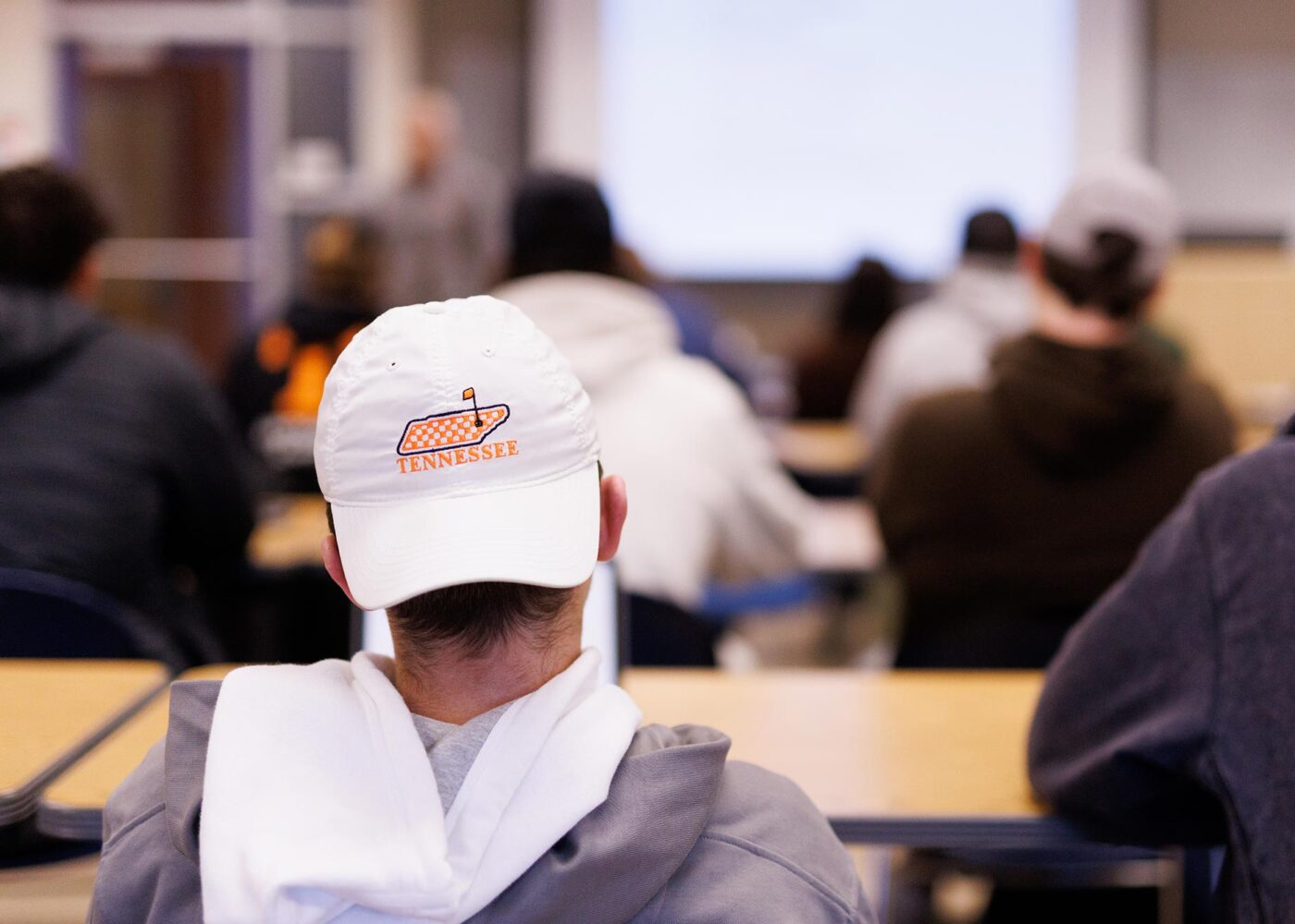 A student sitting in class, wearing a white ball-cap backwards, with a checkered map of Tennessee embroidered on it.