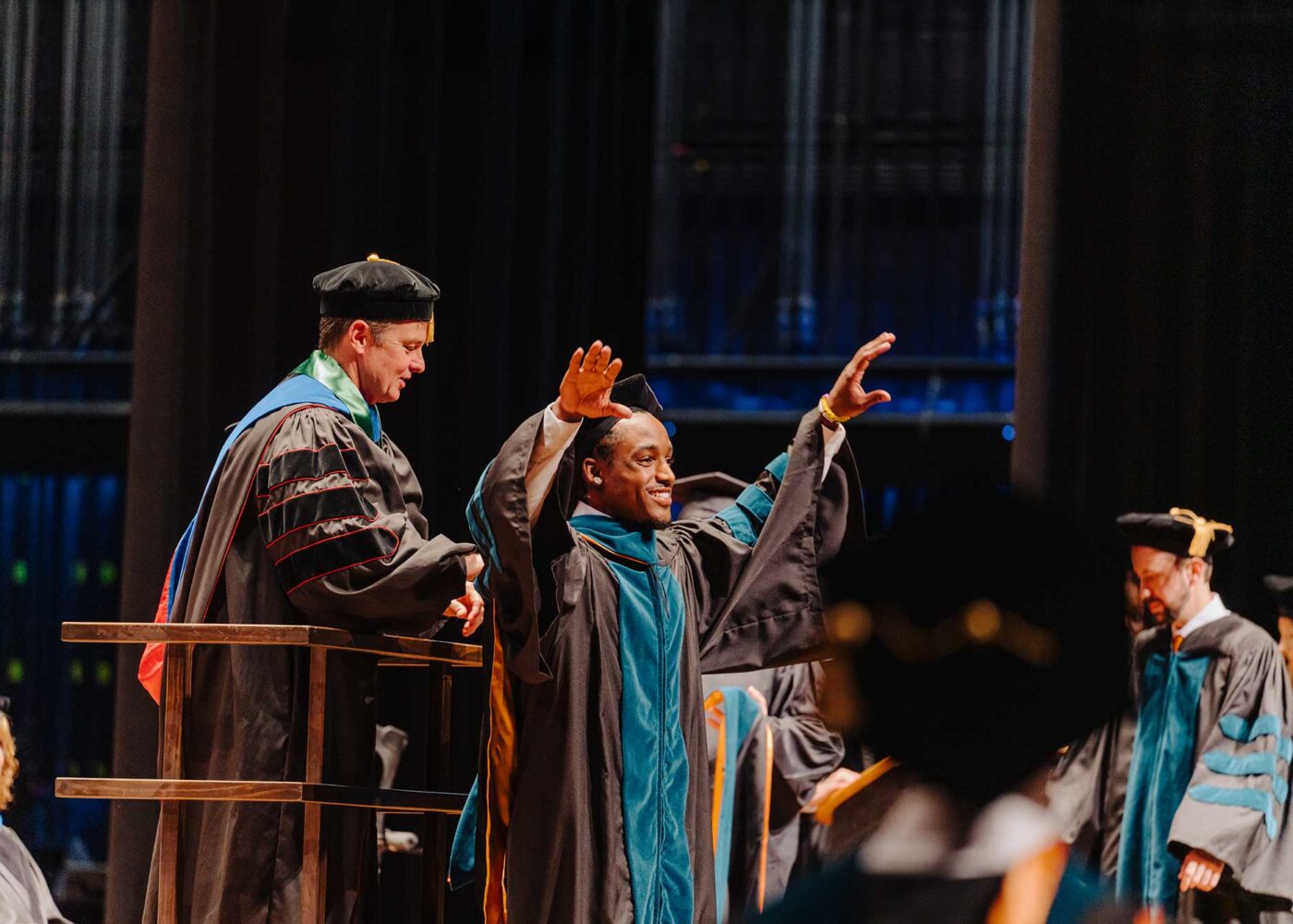 A young man lifts his arm in celebration as his graduation stole is placed on him.