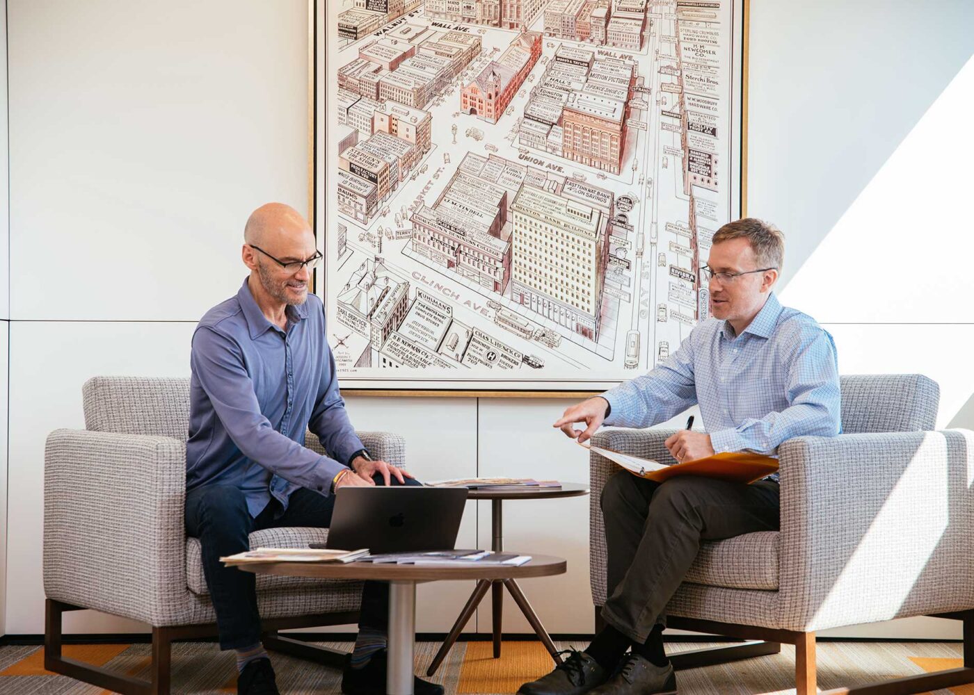 Two men in casual business wear sit in a lounge area while discussing work.
