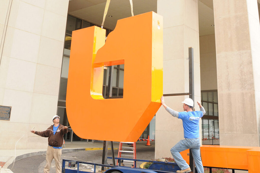 Two construction works work with a crane operator to lift a large metal "U" in preparation to mount it on the side of a building.