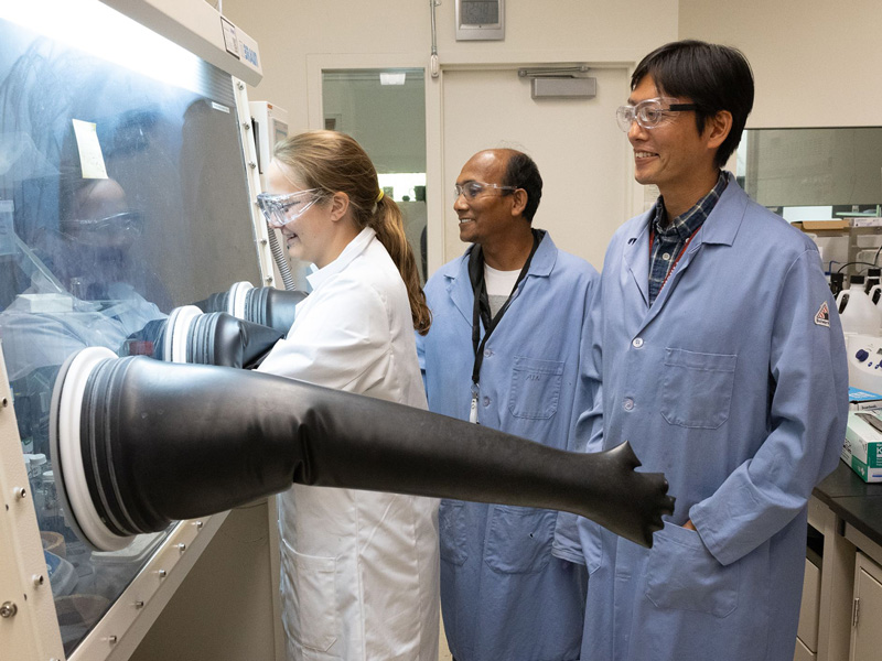 Three scientists wearing lab and safety equipment work on a project using an isolation chamber.