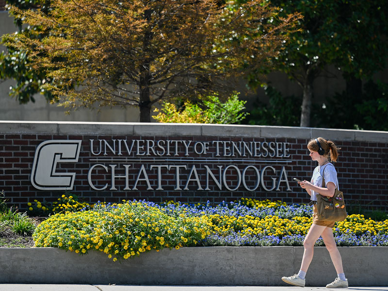A young female student, wearing headphones and using her phone, walking along side a brick wall with the UT Chattanooga sign.