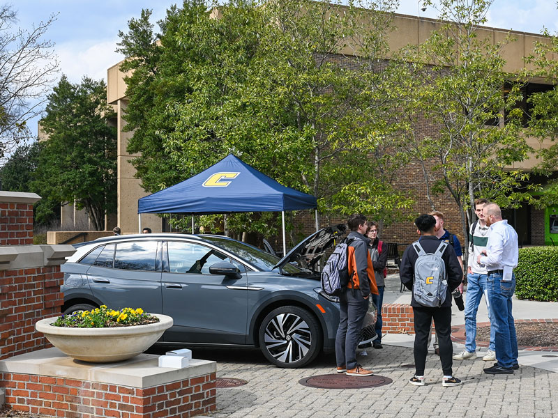 A group of people stand around talking next to a gray vehicle, with the engine hood popped.