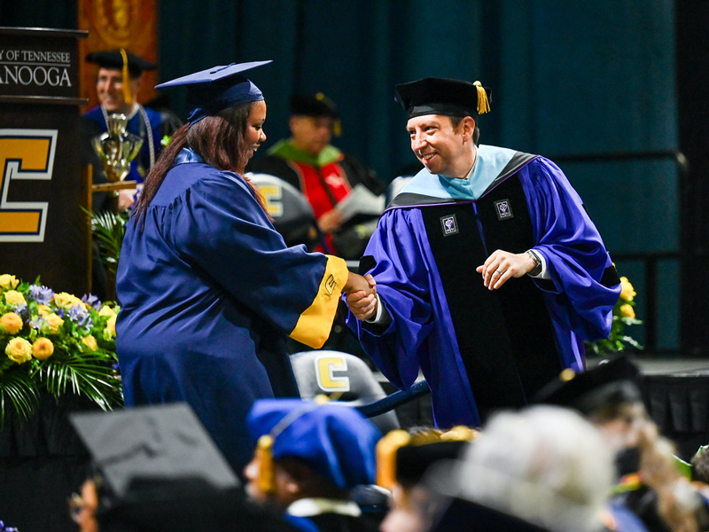 A UTC graduating student shaking Bernie Savarese's hand as she walks across the stage during a graduation ceremony.