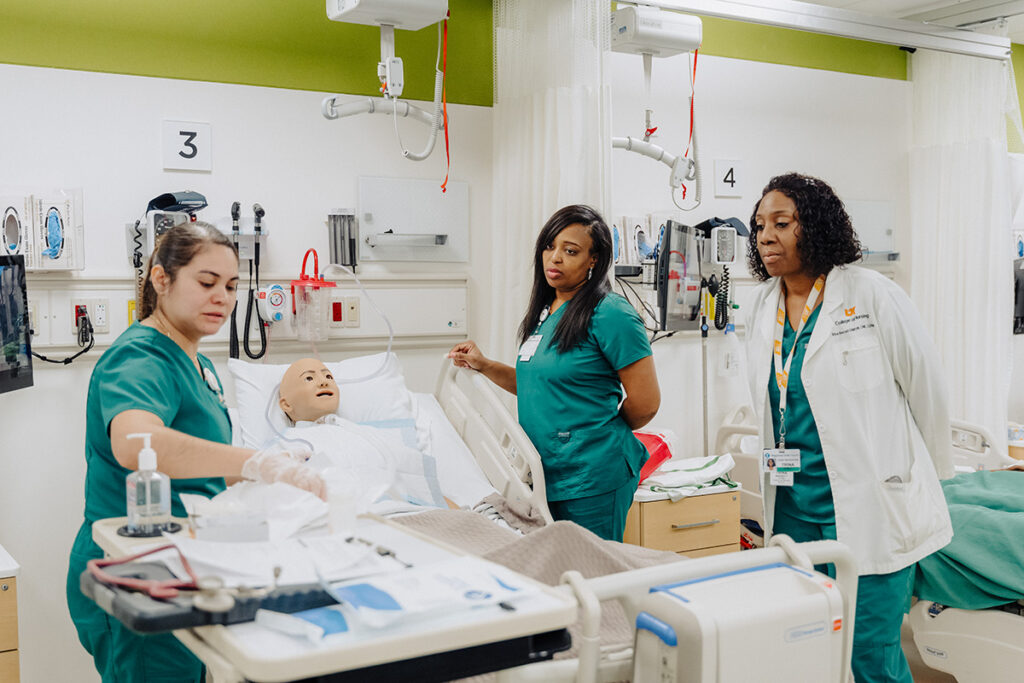Three UT Health Science Center nursing students in green scrubs practice in a simulation lab. One student prepares medical supplies on a cart next to a patient mannequin in a hospital bed, while two other students look on.