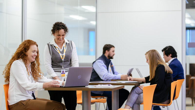 A group of people talking and working on laptops white sitting in a shared break room.