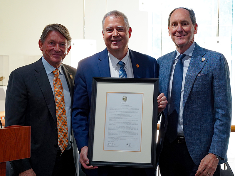 Three men in suits standing for a photo, with the middle man holding up a framed document.