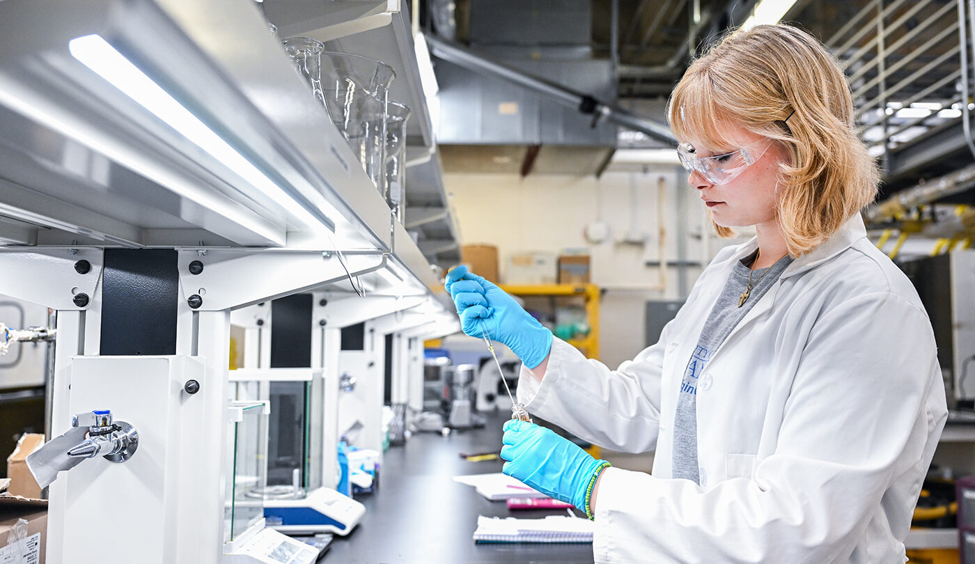 A young woman in a lab coat and safety glasses holds a glass vial in a laboratory. Bright overhead lights illuminate the space filled with various lab equipment.
