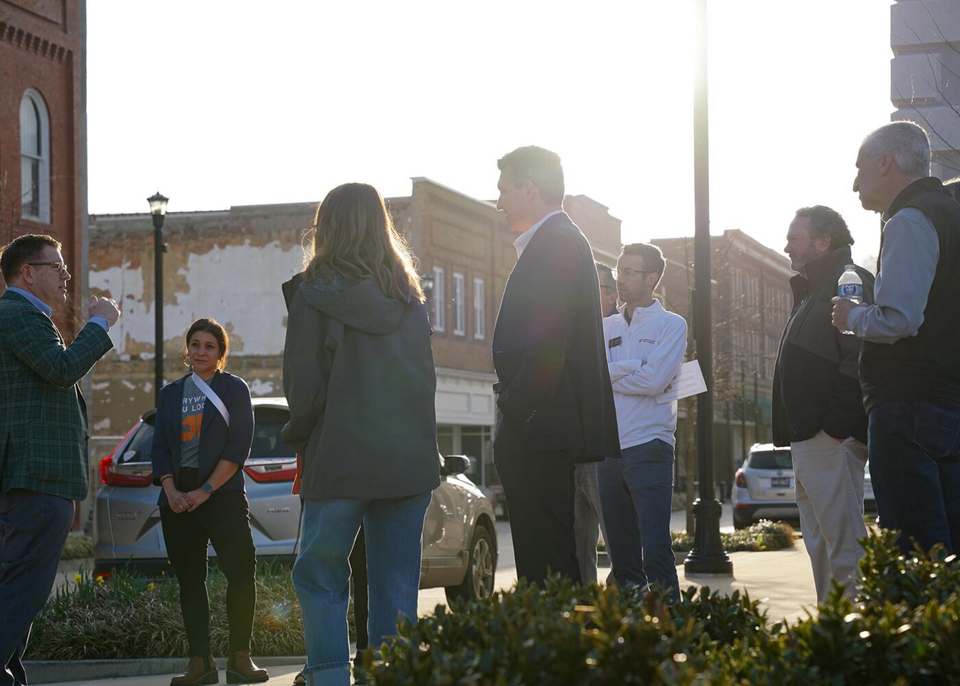 A group of people standing on a down-town sidewalk, listening to a speaker.