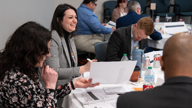 Women and men at an IPS event are sitting at a table and looking at documents.